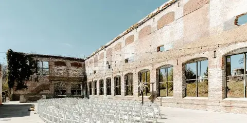 Rows of white chairs set up for a wedding ceremony in front of a historic brick industrial mill building.