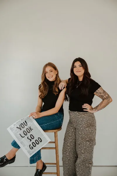 Two women posing together, one holding a newspaper that says 'YOU LOOK SO GOOD'