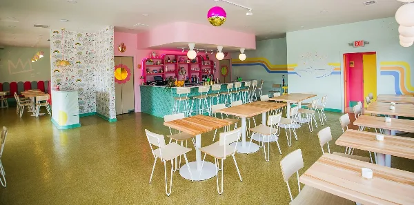 Interior view of a colorful restaurant dining room with wooden tables, white chairs, and a pink bar area.