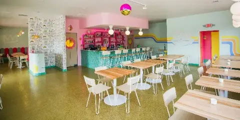 Interior view of a colorful restaurant dining room with wooden tables, white chairs, and a pink bar area.