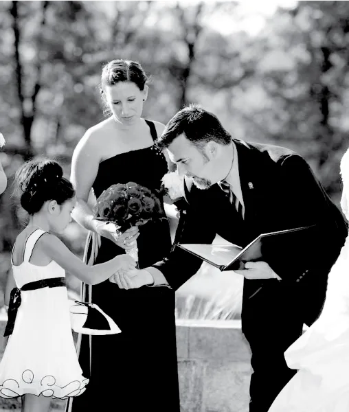 A black and white photo of a man in a suit officiating a wedding ceremony, reading from a folder while a bride and bridesmaid stand nearby.