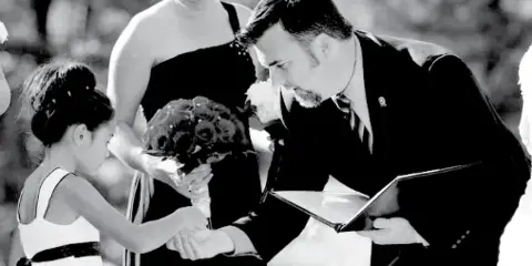 A black and white photo of a man in a suit officiating a wedding ceremony, reading from a folder while a bride and bridesmaid stand nearby.