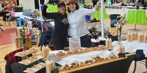 Two women standing behind a catering table filled with appetizers and food displays, with a sign reading 'Alfonso's Catering'.