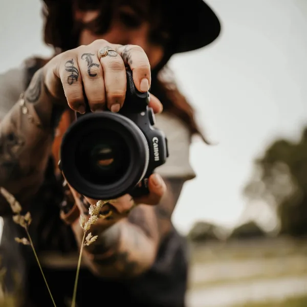 A photographer holding a Canon camera up to the lens, showing tattoos on their hands and a hat.