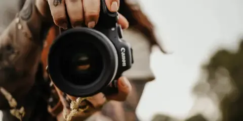A photographer holding a Canon camera up to the lens, showing tattoos on their hands and a hat.
