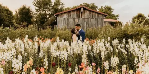 A bride and groom standing in a field of colorful flowers with a weathered wooden barn in the background.