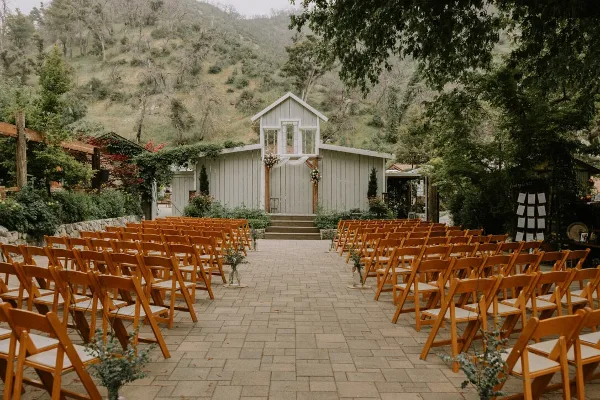 An outdoor wedding ceremony setup featuring rows of wooden chairs leading to a white chapel structure with a wooden arch, set against a backdrop of hills and trees.