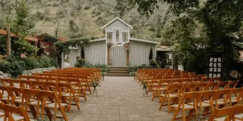 An outdoor wedding ceremony setup featuring rows of wooden chairs leading to a white chapel structure with a wooden arch, set against a backdrop of hills and trees.
