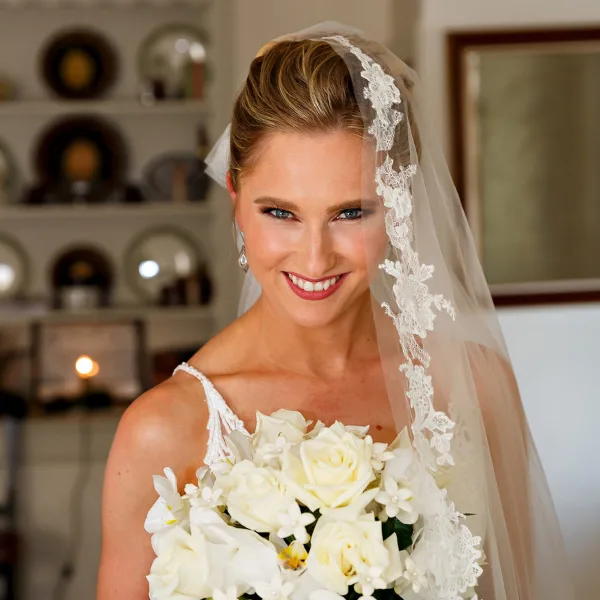 A smiling bride in a white dress and lace veil holding a bouquet of white roses.