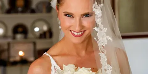 A smiling bride in a white dress and lace veil holding a bouquet of white roses.