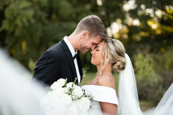 A bride and groom touching foreheads in an outdoor setting with a white bouquet.