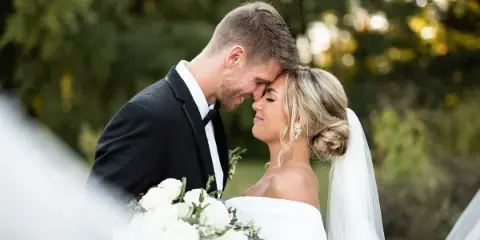 A bride and groom touching foreheads in an outdoor setting with a white bouquet.
