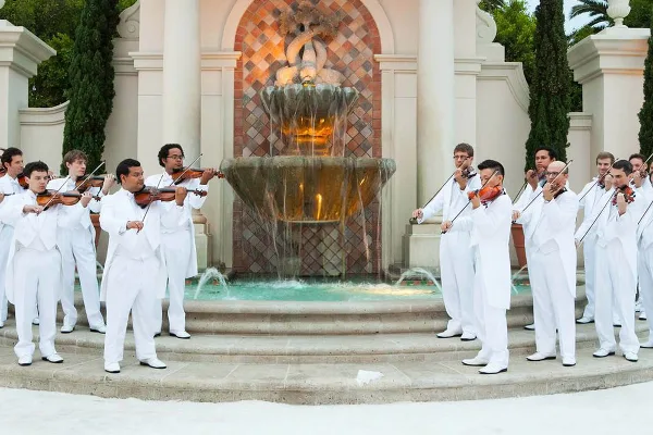 A group of musicians in white suits playing violins in front of a large stone fountain.