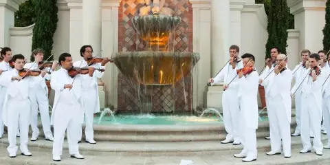 A group of musicians in white suits playing violins in front of a large stone fountain.