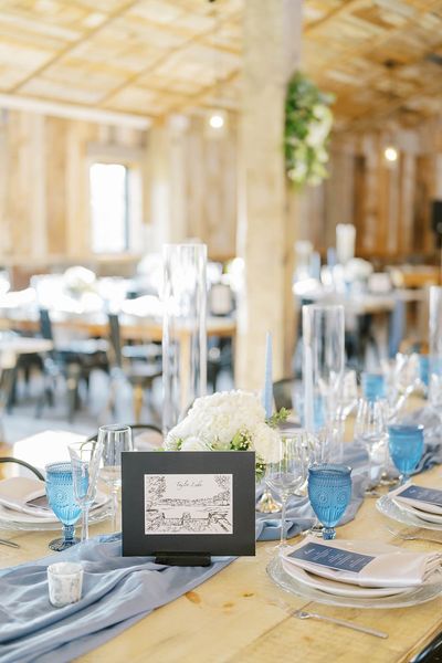 Styled wedding reception table with blue glassware, white floral centerpiece, place cards, and rustic venue interior.