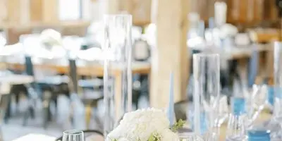 Styled wedding reception table with blue glassware, white floral centerpiece, place cards, and rustic venue interior.