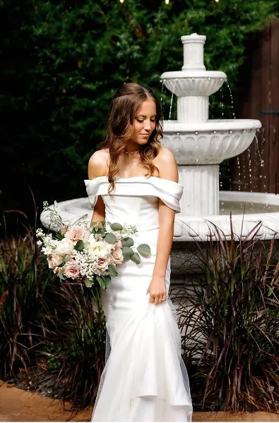 A bride in a white off-the-shoulder wedding dress stands holding a bouquet in front of a white stone fountain and greenery.