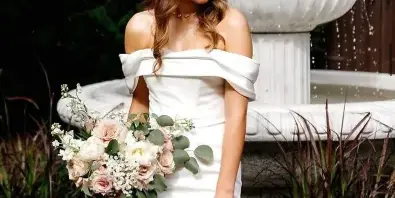 A bride in a white off-the-shoulder wedding dress stands holding a bouquet in front of a white stone fountain and greenery.