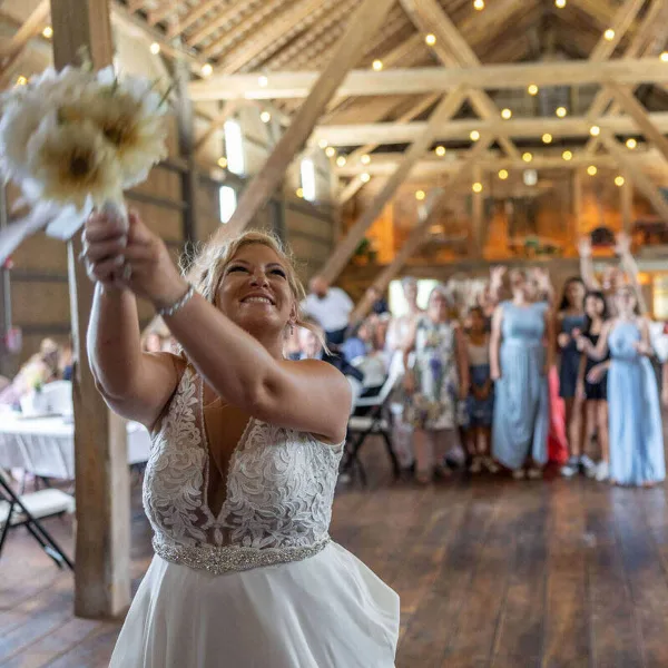 A bride in a white lace dress joyfully tosses her bouquet into a crowd of guests at a rustic wooden venue.