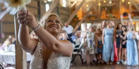 A bride in a white lace dress joyfully tosses her bouquet into a crowd of guests at a rustic wooden venue.