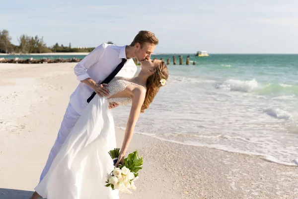 A bride and groom in white wedding attire sharing a kiss on a sandy beach with the ocean in the background.