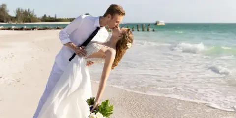 A bride and groom in white wedding attire sharing a kiss on a sandy beach with the ocean in the background.