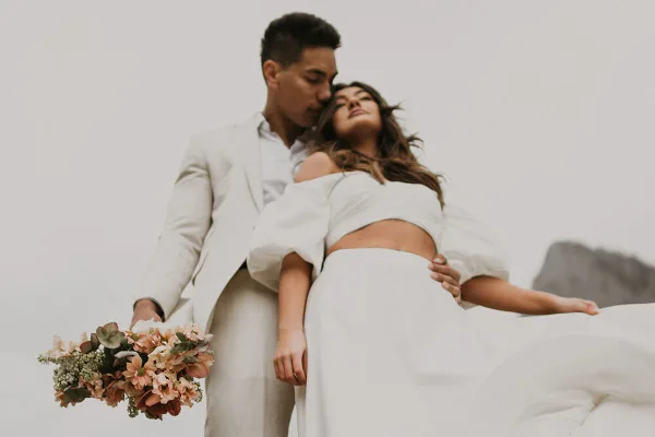 A professional wedding photograph of a couple in white attire, with the groom holding a bouquet of flowers.