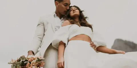 A professional wedding photograph of a couple in white attire, with the groom holding a bouquet of flowers.