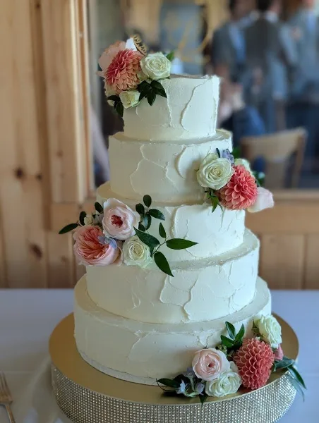A four-tier white wedding cake with textured frosting, decorated with pink and white flowers and greenery, displayed on a gold cake stand.