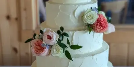 A four-tier white wedding cake with textured frosting, decorated with pink and white flowers and greenery, displayed on a gold cake stand.
