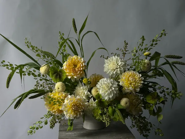 A lush floral arrangement with yellow and white dahlias, greenery, and seed pods in a white vase.