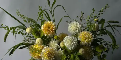 A lush floral arrangement with yellow and white dahlias, greenery, and seed pods in a white vase.