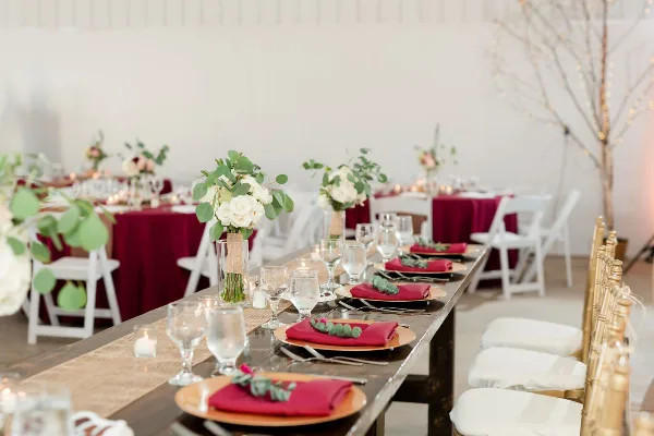A long wooden table set for a wedding reception with red napkins, white floral centerpieces, and glassware.