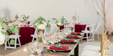 A long wooden table set for a wedding reception with red napkins, white floral centerpieces, and glassware.