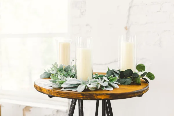 A rustic wooden table stand decorated with greenery and tall candles in glass holders, representing wedding decor styling.