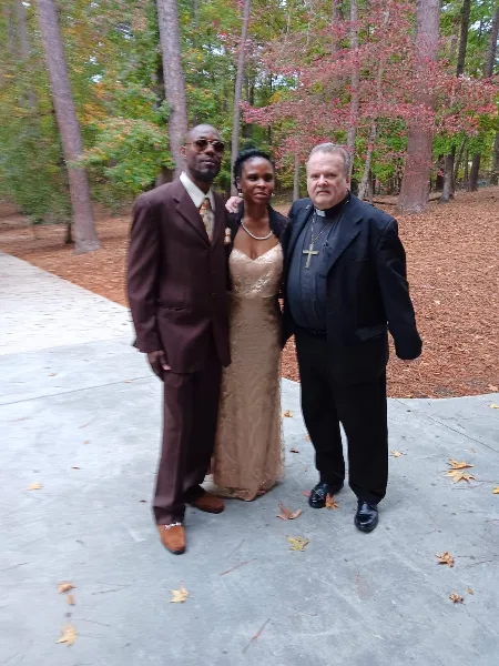A couple in formal attire posing with a clergy member in a wooded outdoor area.