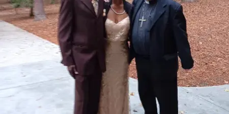 A couple in formal attire posing with a clergy member in a wooded outdoor area.