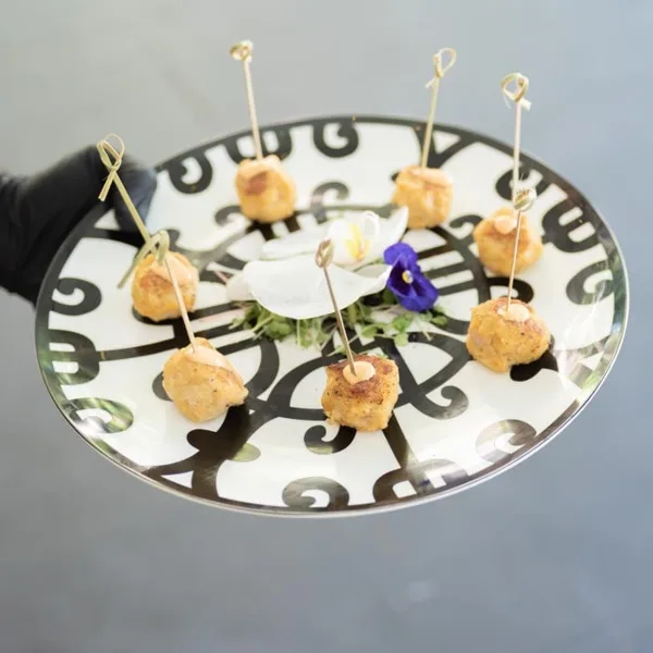 A server in a black glove holding a decorative plate of bite-sized appetizers with toothpicks and edible flowers.
