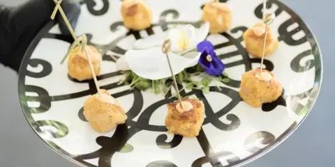 A server in a black glove holding a decorative plate of bite-sized appetizers with toothpicks and edible flowers.