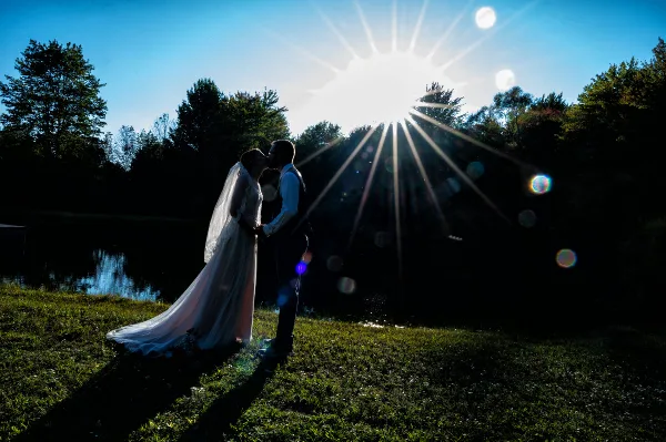 A bride and groom in wedding attire share a kiss on a grassy bank near a pond with a bright sunburst in the background.
