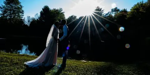 A bride and groom in wedding attire share a kiss on a grassy bank near a pond with a bright sunburst in the background.