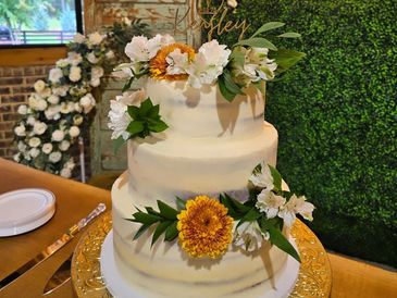 Three-tier white wedding cake decorated with white and yellow flowers and greenery on a gold cake stand.