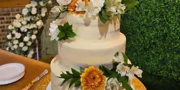 Three-tier white wedding cake decorated with white and yellow flowers and greenery on a gold cake stand.