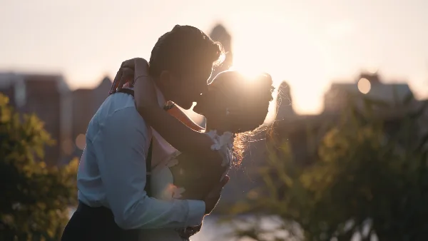 A cinematic, backlit photograph of a bride and groom embracing and kissing at sunset.