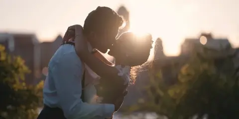 A cinematic, backlit photograph of a bride and groom embracing and kissing at sunset.