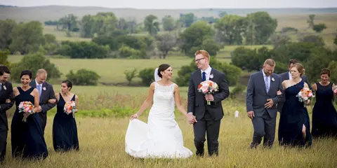 A bride and groom walking down a grassy aisle with their wedding party in a rural field setting.
