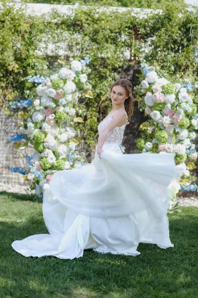 A bride in a white wedding dress standing outdoors in front of a floral arch, showcasing her bridal hair and makeup.