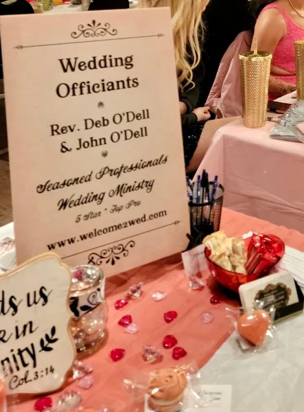 A display table with a sign reading 'Wedding Officiants Rev. Deb O'Dell' and the website welcome2wed.com, decorated with rose petals and a candle.