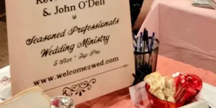 A display table with a sign reading 'Wedding Officiants Rev. Deb O'Dell' and the website welcome2wed.com, decorated with rose petals and a candle.