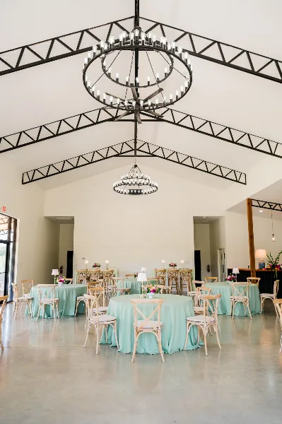 Interior view of a modern barn wedding venue with high ceilings, exposed black trusses, large circular chandeliers, and round tables set with mint green tablecloths.